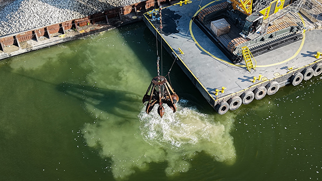 A grapple suspended from a crane releases cobble into the water. The water beneath the grapple is frothy and cloudy from the material disturbing it. In the background is a barge loaded with cobble.